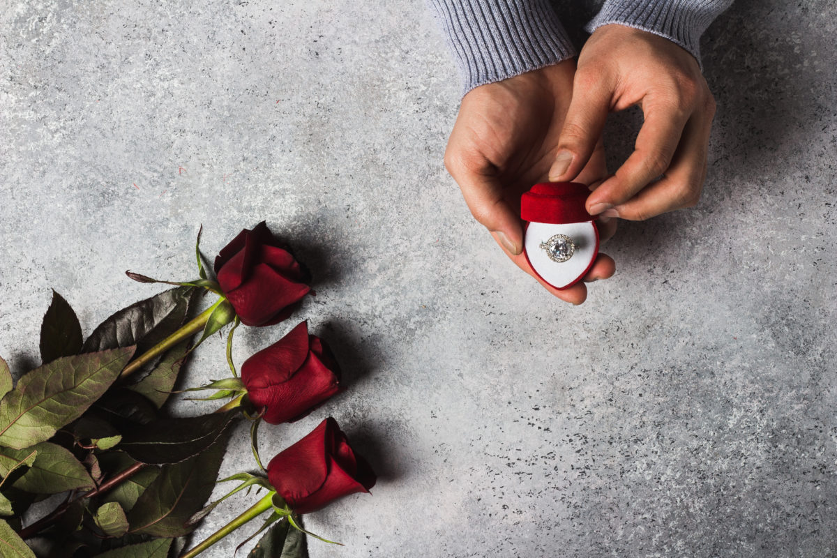 Valentines day romantic man hand holding engagement ring in box marry me wedding with red roses bouquet gift surprise on grey background. Love gift for woman making proposal